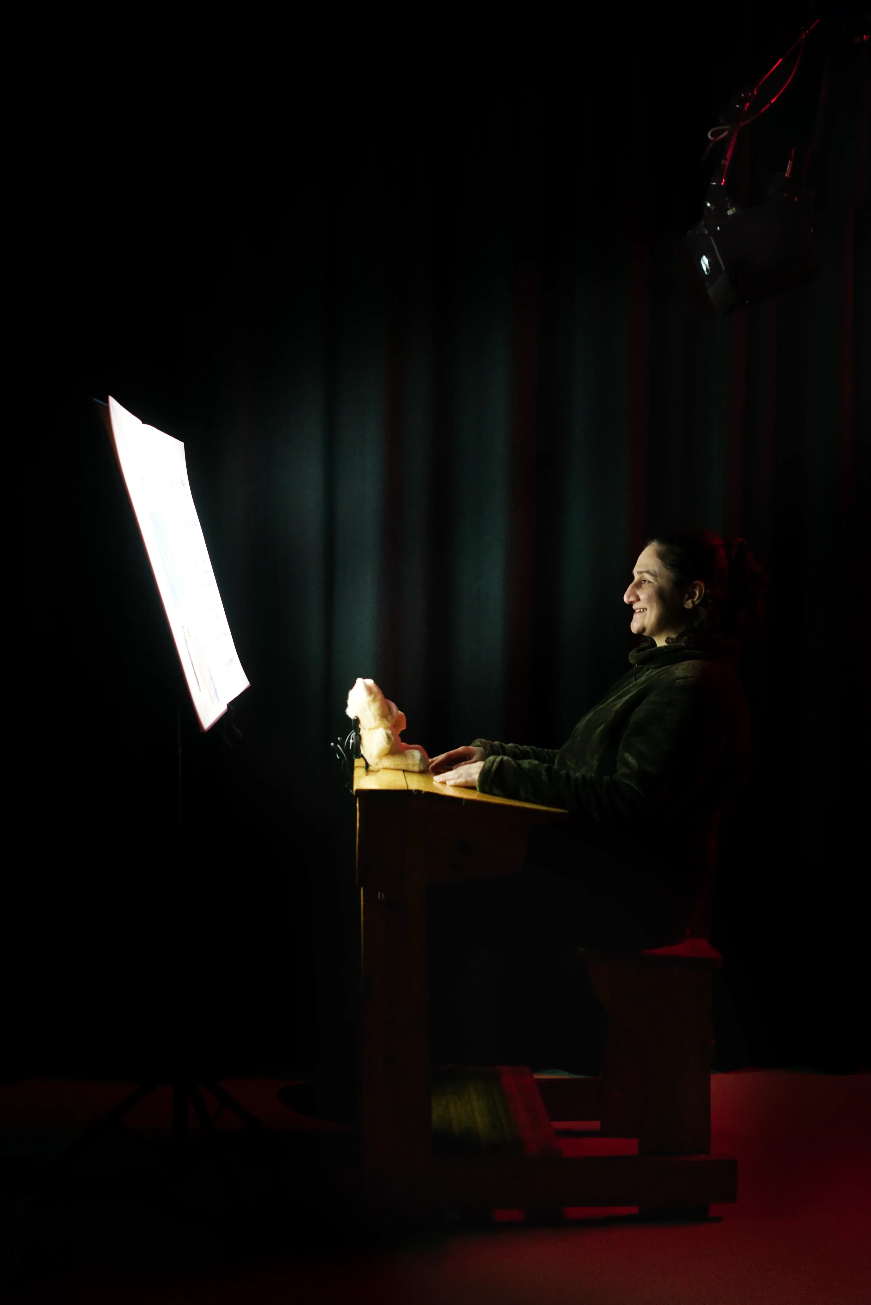 A person smiling in the darkness, sitting at a school desk, staring at a giant book.